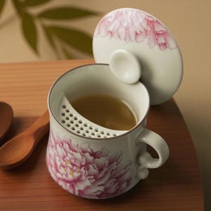 May include: A white ceramic tea cup with a pink peony floral design, a built-in strainer, and a matching lid. The lid features a pink floral pattern. The cup is filled with tea and sits on a wooden surface.