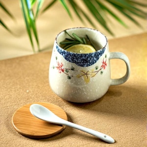 Peut inclure: Une tasse en c&eacute;ramique avec une anse, d&eacute;cor&eacute;e de motifs floraux et d'une bande mouchet&eacute;e bleue. Un dessous de verre en bois et une cuill&egrave;re en c&eacute;ramique blanche sont &agrave; c&ocirc;t&eacute; de la tasse. La tasse contient une tranche de citron et des herbes.
