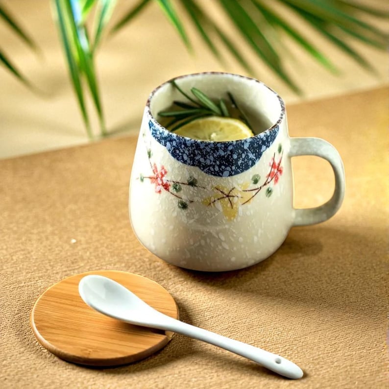 Peut inclure: Une tasse en c&eacute;ramique avec une anse, d&eacute;cor&eacute;e de motifs floraux et d'une bande mouchet&eacute;e bleue. Un dessous de verre en bois et une cuill&egrave;re en c&eacute;ramique blanche sont &agrave; c&ocirc;t&eacute; de la tasse. La tasse contient une tranche de citron et des herbes.