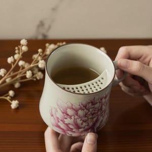 May include: A ceramic tea mug with a built-in strainer and a handle. The mug is decorated with a large pink peony flower design. The mug is filled with tea.