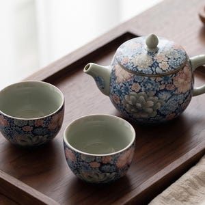Könnte beinhalten: Eine Keramik-Teekanne und zwei passende Teetassen mit einem dichten Blumenmuster in Blau-, Rosa- und Weißtönen. Die Teekanne hat einen runden Körper, einen geschwungenen Ausguss und einen Deckel mit einem kleinen Knauf. Das Innere ist hellgrün.