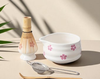 May include: A matcha tea set featuring a white ceramic bowl with pink floral designs, a bamboo whisk, a stainless steel strainer, and a bamboo scoop. The whisk has pink floral accents. The set is designed for preparing matcha tea.