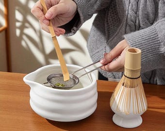 May include: A white ceramic matcha bowl with a spout, a stainless steel strainer, and a bamboo whisk. A bamboo scoop is held over the strainer. A bamboo matcha whisk sits on a wooden surface.