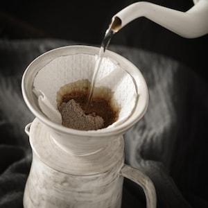 May include: A white ceramic pour-over coffee dripper sits atop a matching ceramic mug. A white gooseneck kettle pours hot water into a white paper filter containing dark brown coffee grounds, creating a brewing process with visible liquid.
