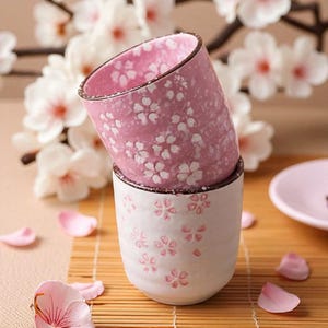 May include: Two ceramic cups stacked, featuring a pink floral design. The top cup is pink with white flowers, while the bottom cup is white with pink flowers. The rims have a dark brown trim. The cups are on a bamboo mat with flower petals.