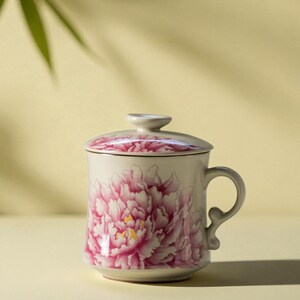 May include: A ceramic mug with a lid, featuring a large pink peony floral design. The mug has a curved handle and a matching floral pattern on the lid. The background is a soft yellow.