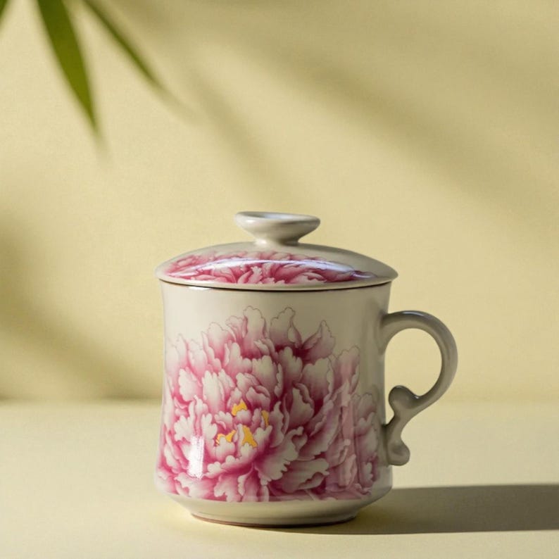 May include: A ceramic mug with a lid, featuring a large pink peony floral design. The mug has a curved handle and a matching floral pattern on the lid. The background is a soft yellow.