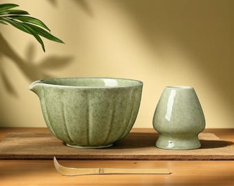 May include: A matcha tea set featuring a light green ceramic bowl with a spout, a small matching whisk holder, and a bamboo whisk. The set is displayed on a wooden surface, ideal for preparing traditional matcha.