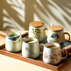 Peut inclure: Plusieurs tasses en c&eacute;ramique avec des motifs peints &agrave; la main, notamment floraux, de poissons et abstraits. Certaines tasses ont des couvercles en bois. Les tasses sont pr&eacute;sent&eacute;es sur un plateau en bois, mettant en valeur une esth&eacute;tique rustique.