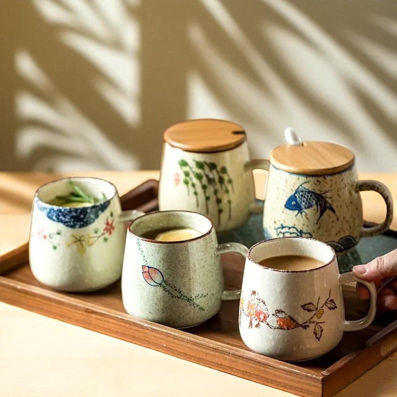 Peut inclure: Plusieurs tasses en c&eacute;ramique avec des motifs peints &agrave; la main, notamment floraux, de poissons et abstraits. Certaines tasses ont des couvercles en bois. Les tasses sont pr&eacute;sent&eacute;es sur un plateau en bois, mettant en valeur une esth&eacute;tique rustique.