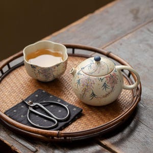 May include: A ceramic teapot and a matching flower-shaped teacup, both featuring a cream-coloured base with hand-painted purple and green floral patterns. A small pair of dark metal scissors rests on a black fabric square with white dots.