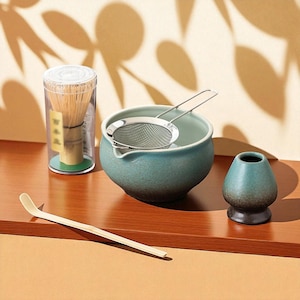 Może przedstawiać: Zestaw do herbaty matcha, w tym bambusowa miotełka w przezroczystym pojemniku, turkusowa ceramiczna miska ze stalowym sitkiem, mała turkusowa ceramiczna filiżanka do herbaty i bambusowa łyżka. Zestaw jest ułożony na drewnianej powierzchni.