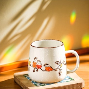 Peut inclure: Une tasse en c&eacute;ramique blanche avec un bord et une anse marron. La tasse pr&eacute;sente un motif peint &agrave; la main de trois oiseaux perch&eacute;s sur une branche avec des couleurs orange et marron. La tasse est pos&eacute;e sur un dessous de verre en bois.