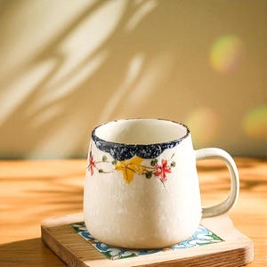 Peut inclure: Une tasse en c&eacute;ramique blanche avec une forme arrondie et une anse. Le bord est d&eacute;cor&eacute; d'un motif bleu fonc&eacute; et blanc, et le corps pr&eacute;sente des motifs floraux rouges, jaunes et verts. La tasse repose sur un dessous de verre en bois.