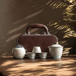 Könnte beinhalten: Ein Keramik-Teeservice mit floralen Designs, darunter eine Teekanne, kleine Tassen und ein Behälter mit Deckel, wird vor einem braunen Lederkoffer präsentiert. Das Teeservice ist weiß mit rosa Blumenmustern. Der Koffer hat einen braunen Griff.