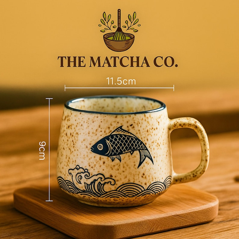 Peut inclure: Tasse en c&eacute;ramique tachet&eacute;e avec un motif de poisson. La tasse beige pr&eacute;sente une illustration de poisson bleu et un motif de vagues. Elle mesure 9 cm de haut et 11,5 cm de large. Le texte "THE MATCHA CO." est au-dessus.