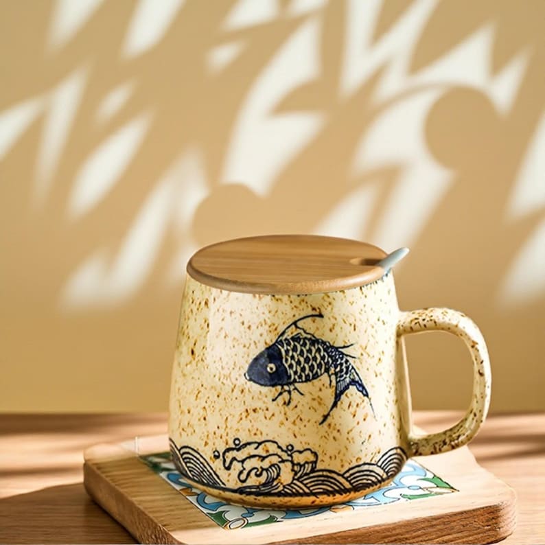 Peut inclure: Une tasse en c&eacute;ramique avec un motif de poisson, un couvercle en bois et une petite cuill&egrave;re. La tasse est beige avec une illustration de poisson bleu et un motif de vagues. La poign&eacute;e est de couleur beige assortie. La tasse repose sur un dessous de verre en bois.