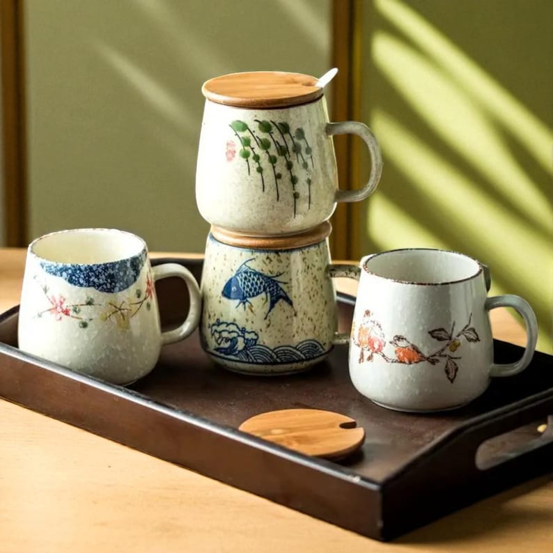 Peut inclure: Quatre mugs en c&eacute;ramique avec des poign&eacute;es, chacun avec des motifs peints &agrave; la main uniques. Un mug a un couvercle et une cuill&egrave;re. Les mugs sont d&eacute;cor&eacute;s de motifs floraux, de poissons et d'oiseaux en bleu, rouge et vert. Ils sont sur un plateau en bois brun fonc&eacute;.