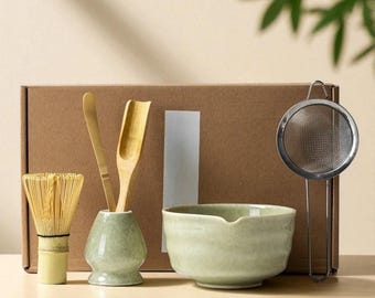 May include: A matcha tea set with a bamboo whisk, scoop, and spoon, a ceramic bowl, a strainer, and a small vase. The items are light green and beige, arranged in front of a brown box.