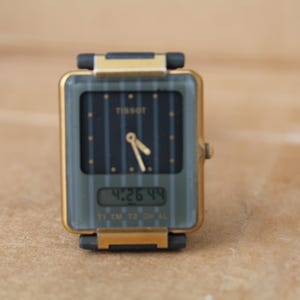 Puede incluir: Reloj digital Tissot vintage con esfera rectangular. El reloj tiene una caja dorada y una esfera gris oscura con líneas verticales y el logotipo de Tissot. La pantalla digital muestra la hora: 4:26:44.