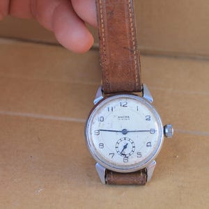 May include: A vintage wristwatch with a round silver-tone metal case and a worn brown leather strap. The off-white dial features black Arabic numerals, a small sub-dial for seconds at the six o'clock position, and the text Ancre 15 Rubis.