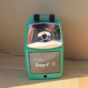 May include: A green Carl Angel-5 pencil sharpener with a metallic silver front plate and two black plastic tabs at the top. The front features a clear plastic shavings receptacle with the brand name Carl and the model name Angel-5 printed on it.