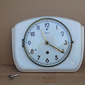 May include: A white wall clock with a rectangular ceramic body and a circular face featuring black Arabic numerals, gold-toned hands, and the Diehl brand name. A small metal winding key is positioned separately below the clock unit.