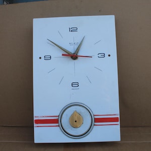 May include: A rectangular white wall clock featuring black numerals at the 3, 6, 9, and 12 positions. The clock has gold-toned hour and minute hands, a red second hand, and a circular gold-toned pendulum element above two horizontal red stripes.