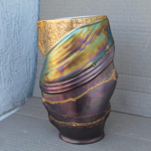 Puede incluir: Un jarrón cilíndrico con una forma orgánica irregular. La sección superior presenta un acabado dorado texturizado, mientras que la parte central muestra un brillo metálico iridiscente que cambia entre verde, azul y morado. La base es morado oscuro.