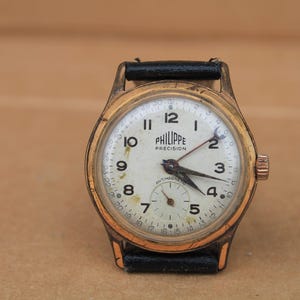 May include: A vintage wristwatch with a gold-tone metal case and a black leather strap. The off-white dial features black Arabic numerals, the text PHILIPPE PRECISION, a small sub-dial at the six o'clock position, and an outer date ring.