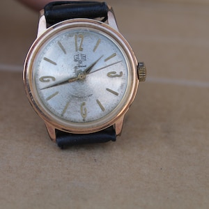 Könnte beinhalten: Vintage goldfarbene Armbanduhr mit silbernem Zifferblatt und schwarzem Lederarmband. Das Zifferblatt der Uhr hat goldfarbene Stundenmarkierungen und Zeiger, mit der Zahl 12 oben. Die Uhr hat eine kleine Krone und die Marke "GUB" ist sichtbar.