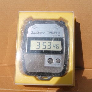 May include: A black and silver digital Hanahart TIMOPHIL stopwatch, displaying the time 3:53:46. The stopwatch is encased in a clear plastic box with a yellow base. The stopwatch has two buttons and a split function.