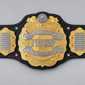 Könnte beinhalten: Ein schwarzer Leder-Meisterschaftsgürtel mit einer großen zentralen Gold- und Silberplatte mit der Aufschrift IWGP HEAVYWEIGHT CHAMPION. Der Gürtel enthält vier kleinere Seitenplatten und goldene Metallnieten an den Enden.