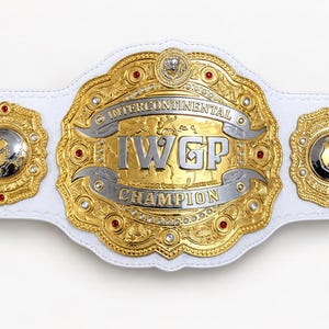 Könnte beinhalten: Ein weißer Leder-Meisterschaftsgürtel mit einer großen zentralen Gold- und Silberplatte mit der Aufschrift IWGP INTERCONTINENTAL CHAMPION. Vier kleinere Goldplatten mit silbernen Globus-Einlagen und roten Akzenten.