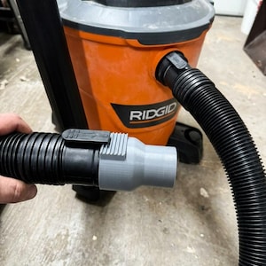 May include: An orange and black RIDGID wet/dry vacuum with a black hose and a gray adapter. The hose is connected to the vacuum and held by a hand. The vacuum has the RIDGID logo on the front.