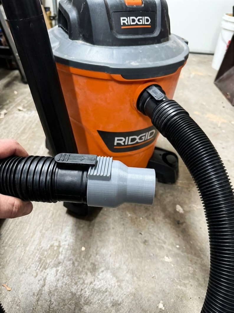May include: An orange and black RIDGID wet/dry vacuum with a black hose and a gray adapter. The hose is connected to the vacuum and held by a hand. The vacuum has the RIDGID logo on the front.