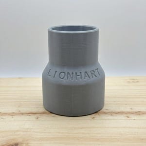 May include: A gray plastic pipe fitting with the word "LIONHART" embossed on the side. The fitting has a wider base and a narrower top. It is sitting on a light-colored wooden surface.