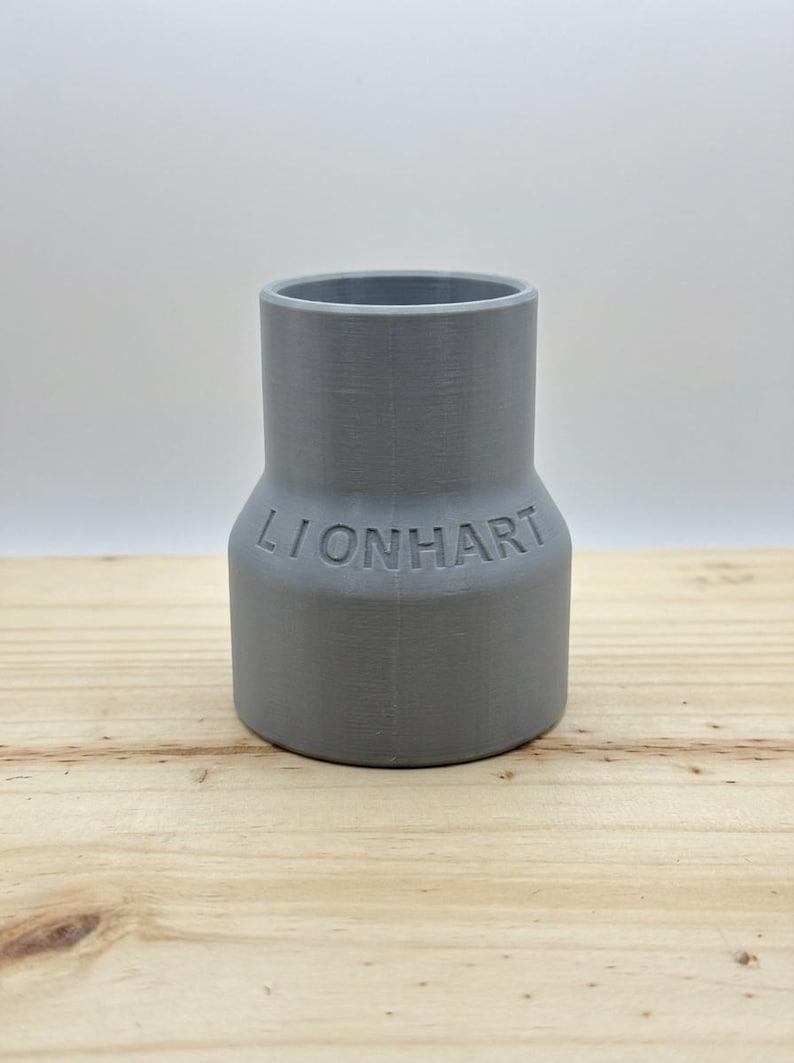 May include: A gray plastic pipe fitting with the word "LIONHART" embossed on the side. The fitting has a wider base and a narrower top. It is sitting on a light-colored wooden surface.