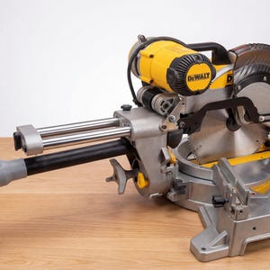 May include: A yellow and silver miter saw with a black handle and a circular saw blade. The saw has a black hose attached to it and is sitting on a wooden surface. The brand name "DEWALT" is visible on the motor housing.