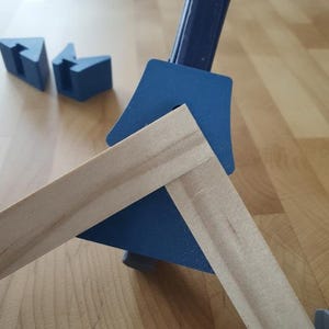 May include: A blue corner clamp holding two wooden frame pieces together. Two additional blue corner pieces are visible in the background. The wood has a light brown color, and the clamp is on a light brown wooden surface.