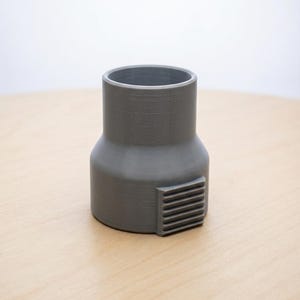 May include: A gray, cylindrical adapter with a rectangular vent detail. The adapter has a wider base and a narrower top, and it sits on a light-colored wooden surface. The background is a plain white.