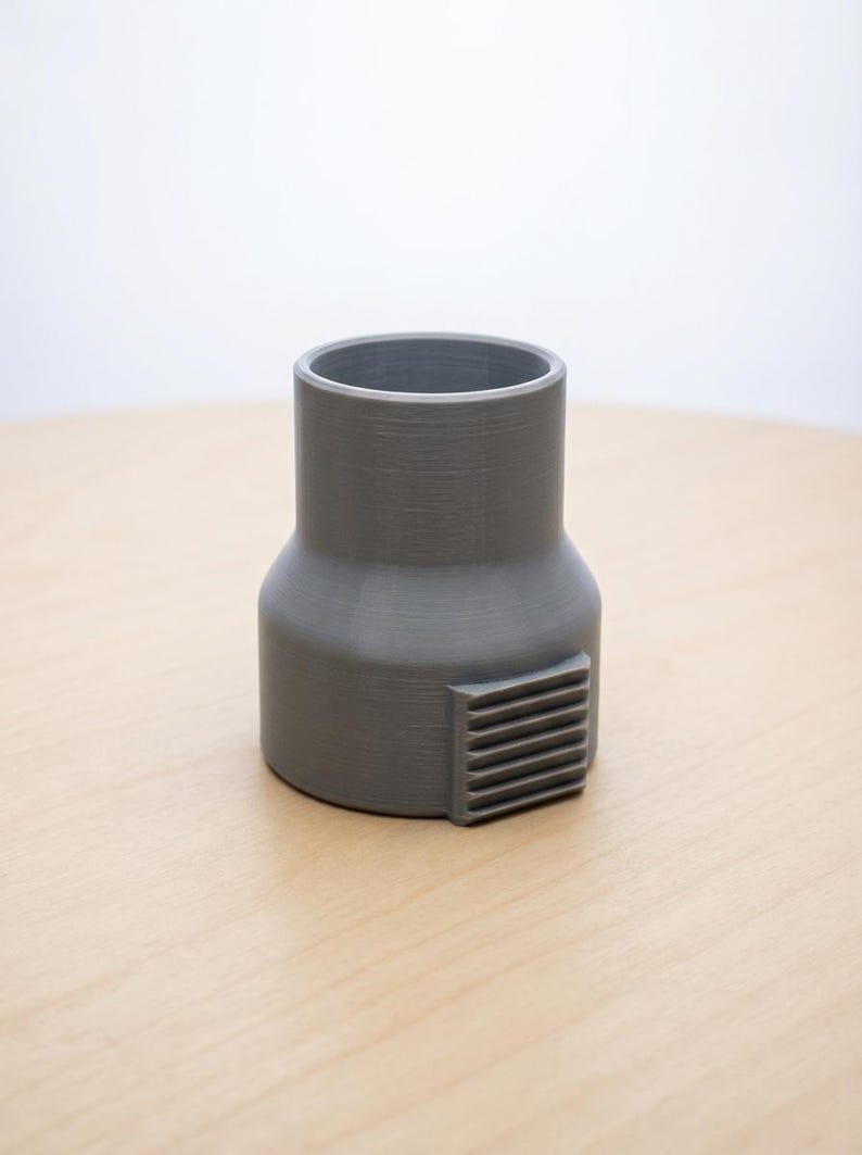 May include: A gray, cylindrical adapter with a rectangular vent detail. The adapter has a wider base and a narrower top, and it sits on a light-colored wooden surface. The background is a plain white.