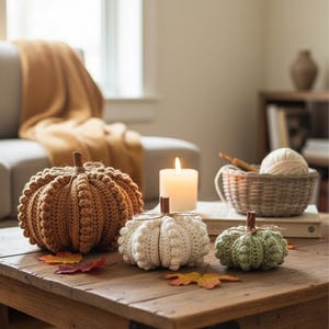 May include: Three handmade crochet pumpkins in brown, cream, and green sit on a wooden table. A lit candle and autumn leaves enhance the seasonal display. A basket with yarn and knitting needles is also visible.