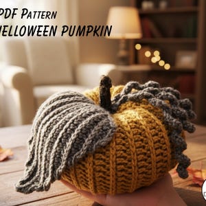 May include: A handmade, crocheted pumpkin in shades of yellow and gray, with the text "PDF PATTERN HELLOWEEN PUMPKIN" at the top. The pumpkin is held in a person's hand, with a wooden table and fall leaves in the background.