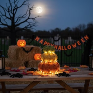 May include: An outdoor Halloween display shows a stack of three crocheted pumpkins in orange, yellow, and green, lit by fairy lights. The scene includes a "Happy Halloween" banner, hay bales, and decorative spiders, set under a full moon in a night sky.