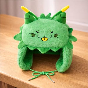 May include: A plush green hat shaped like a cartoon creature with a smiling face, yellow tongue, and antennae. The hat has ear flaps and a green tie. The hat is made of soft, fuzzy material.