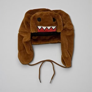 May include: A brown plush hat with a cartoon face design. The hat features a wide-open mouth with white teeth and a red tongue, black eyes, and long ear flaps. A brown drawstring is attached.
