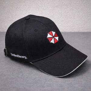 Puede incluir: Gorra de béisbol negra con ribete blanco en la visera. La gorra presenta un logotipo de paraguas rojo y blanco en la parte delantera y el texto "Umbrellacorp." en el lateral. Estilo clásico.