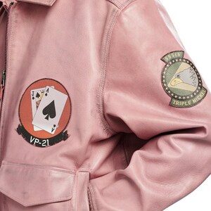Pu&ograve; includere: Un giubbotto bomber in pelle rosa chiaro con una toppa circolare sul petto raffigurante due carte da gioco e il testo VP-21, e una toppa circolare sulla manica con un'aquila e il testo 555th TRIPLE NICKEL.