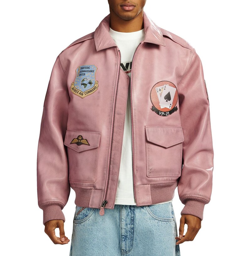 Pu&ograve; includere: Un giubbotto bomber in pelle rosa polvere con cerniera frontale, polsini a coste e due tasche con patta. Il petto presenta toppe ricamate, tra cui un emblema Strategic Reconnaissance Center Avirex Air Command e una grafica di carte da gioco VP-21.